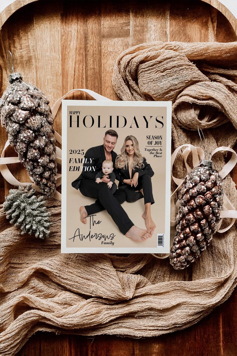 May include: A magazine cover with the text "HAPPY HOLIDAYS 2025 FAMILY EDITION" featuring a family portrait. The magazine is surrounded by decorative pinecone ornaments and draped fabric, creating a festive holiday theme.