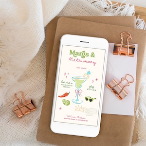 Op de afbeelding: Een witte smartphone toont een kleurrijke uitnodiging voor een vrijgezellenweekend. De uitnodiging luidt "Margs & Matrimony" met een margarita-illustratie, zonnebril en andere feestelijke graphics. Koperen paperclips en briefpapier zijn ook zichtbaar.