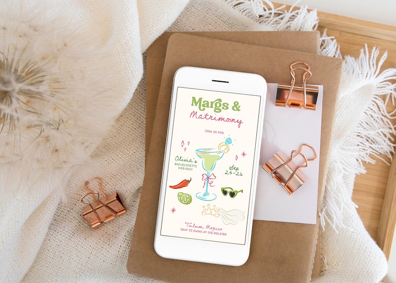 Op de afbeelding: Een witte smartphone toont een kleurrijke uitnodiging voor een vrijgezellenweekend. De uitnodiging luidt "Margs & Matrimony" met een margarita-illustratie, zonnebril en andere feestelijke graphics. Koperen paperclips en briefpapier zijn ook zichtbaar.