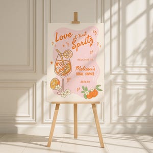 以下が含まれることがあります： 木製のイーゼルに「Love at First Spritz」というオレンジ色の文字が書かれた看板。オレンジのスライスが入ったカクテルのイラストと「Welcome to Melissa's Bridal Shower 26.06.XX」というテキストが特徴です。