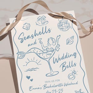 May include: A light cream-colored invitation with blue line art. The text reads "Seashells and Wedding Bells" with illustrations of a mermaid in a martini glass, seashells, and a disco ball. A gold ribbon is draped across the top.