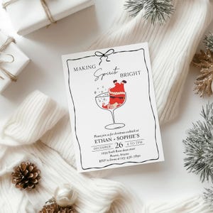 May include: A white invitation card with a black border and the words "MAKING Spirit BRIGHT". The card features a Santa Claus illustration in a cocktail glass. The invitation is for Christmas cocktails at Ethan + Sophie's on December 26.