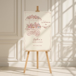 May include: A cream-colored sign on a wooden easel, featuring a floral cart illustration in a reddish-brown hue. The sign reads "Welcome to Sophie's Bridal Shower" with the date below. The easel is made of light-colored wood.