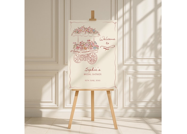 May include: A cream-colored sign on a wooden easel, featuring a floral cart illustration in a reddish-brown hue. The sign reads "Welcome to Sophie's Bridal Shower" with the date below. The easel is made of light-colored wood.