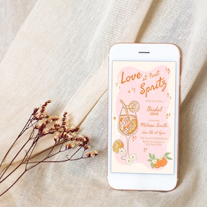 May include: A smartphone displays a bridal shower invitation with the text "Love at First Spritz." The invitation features an illustration of a cocktail, oranges, and flowers. The phone rests on a beige fabric with dried floral accents.