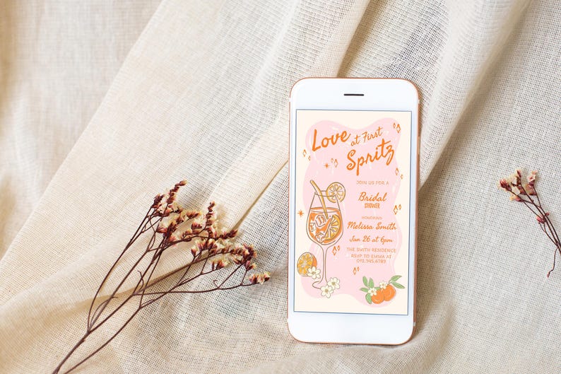 May include: A smartphone displays a bridal shower invitation with the text "Love at First Spritz." The invitation features an illustration of a cocktail, oranges, and flowers. The phone rests on a beige fabric with dried floral accents.