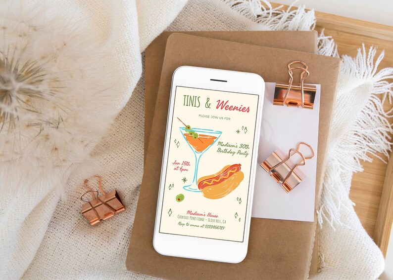 May include: A smartphone displays a party invitation with the text "Tinis & Weenies." The invitation features a cocktail and hot dog illustration. The phone rests on a stack of brown paper with copper-colored binder clips.