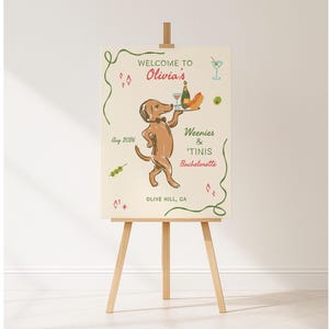 May include: A rectangular welcome sign on a wooden easel featuring a brown dachshund wearing a bowtie and carrying a tray with a bottle and hot dog. Text reads: WELCOME TO Olivia's, Weenies & 'Tinis Bachelorette, Aug 2026, and OLIVE HILL, CA.