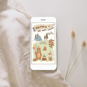 Puede incluir: Un smartphone muestra una invitaci&oacute;n de cumplea&ntilde;os con tem&aacute;tica de camping. La ilustraci&oacute;n presenta un oso con globos, un zorro, una tienda de campa&ntilde;a y una fogata. El texto dice "Mathew is One Happy Camper" con detalles del evento. El tel&eacute;fono est&aacute; sobre una superficie neutra.