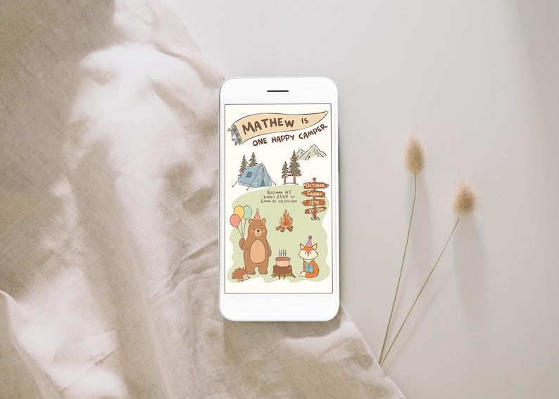 Puede incluir: Un smartphone muestra una invitaci&oacute;n de cumplea&ntilde;os con tem&aacute;tica de camping. La ilustraci&oacute;n presenta un oso con globos, un zorro, una tienda de campa&ntilde;a y una fogata. El texto dice "Mathew is One Happy Camper" con detalles del evento. El tel&eacute;fono est&aacute; sobre una superficie neutra.