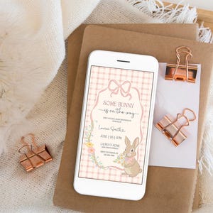 Op de afbeelding: Een smartphone toont een uitnodiging voor een babyshower met de tekst "Some Bunny is on the Way". De uitnodiging heeft een konijnenillustratie, bloemaccenten en een roze ruitjesachtergrond. Koperkleurige paperclips en briefpapier zijn ook zichtbaar.