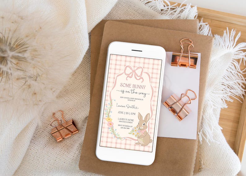 Op de afbeelding: Een smartphone toont een uitnodiging voor een babyshower met de tekst "Some Bunny is on the Way". De uitnodiging heeft een konijnenillustratie, bloemaccenten en een roze ruitjesachtergrond. Koperkleurige paperclips en briefpapier zijn ook zichtbaar.