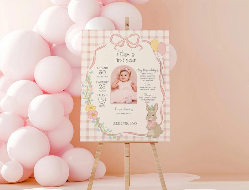 May include: A pink and white decorative sign on an easel, featuring a baby's first-year details. The sign includes a photo, measurements, and a bunny illustration. The sign is surrounded by pink balloons.