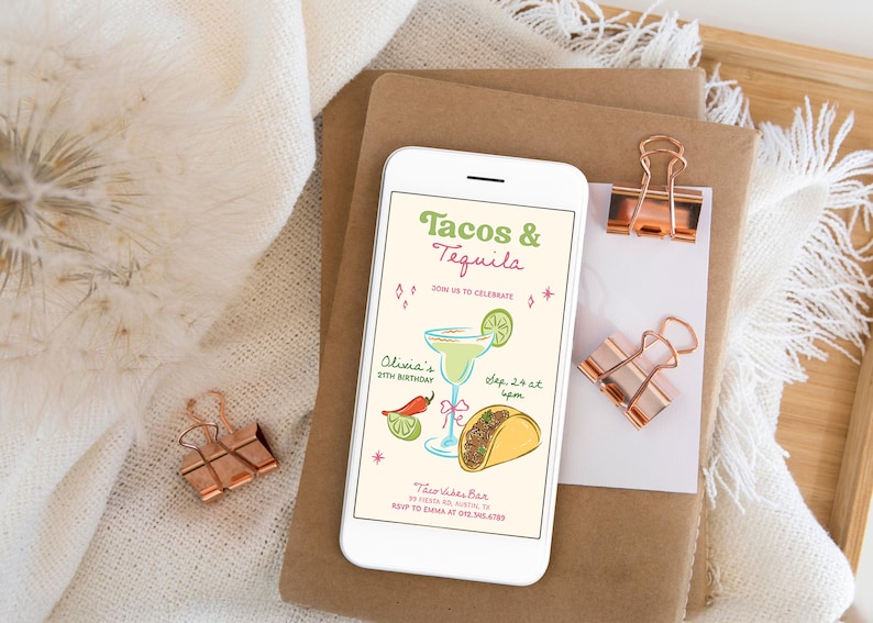May include: A smartphone displays a party invitation with the text "Tacos & Tequila." The invitation features illustrations of a margarita, a taco, and a chili pepper. The phone rests on a stack of brown paper with rose gold binder clips.