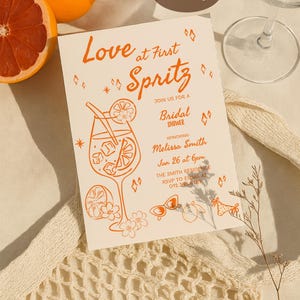 May include: A bridal shower invitation with orange text and illustrations, featuring a cocktail glass, flowers, and sunglasses. The invitation reads "Love at First Spritz" and includes event details. Oranges and a wine glass are also visible.