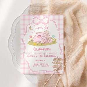 May include: A pink and white invitation card with a bow design at the top. The card features a tent illustration with the words "Let's Go Glamping!" and details for Emily's 7th birthday. The card is on a white plate with a beige fabric.