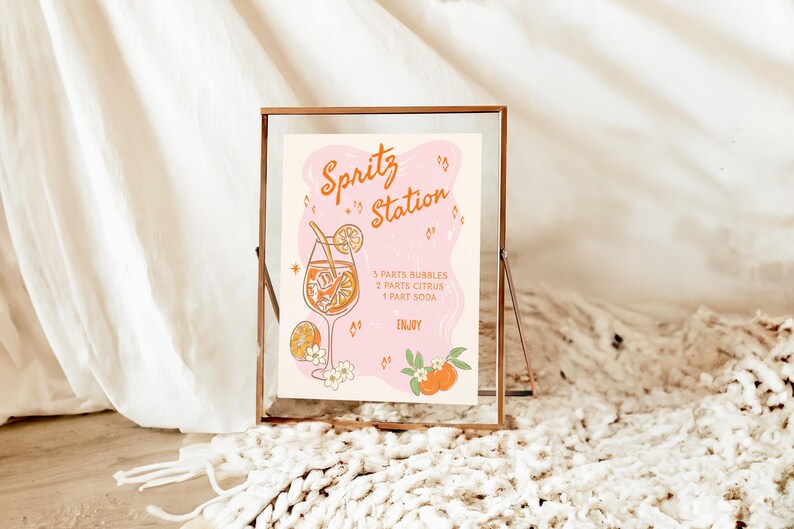 May include: A framed print with the text "Spritz Station" in orange script. The illustration features a cocktail glass with orange slices and a recipe. The background is a soft pink shape with orange accents and floral details.