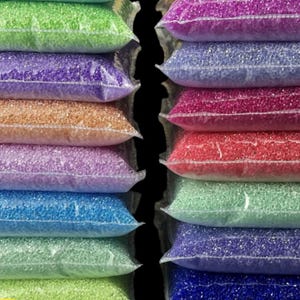 May include: A close-up shot of several clear plastic bags filled with colorful beads. The bags are stacked, showcasing a variety of colors including green, purple, orange, pink, blue, and red. The beads appear to be small and uniform in size.