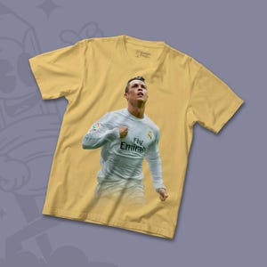 May include: A yellow t-shirt featuring a printed image of a football player wearing a white jersey with the text "Fly Emirates" on the front. The player is raising his right arm in the air.