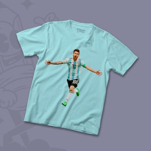 Peut inclure: Un t-shirt bleu clair avec un motif repr&eacute;sentant un joueur de football portant un maillot bleu et blanc avec le num&eacute;ro 10. Le joueur est en l'air, les bras tendus, c&eacute;l&eacute;brant un but.
