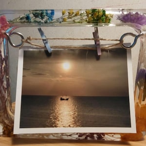 Könnte beinhalten: Ein klarer Bilderrahmen aus Harz mit eingebetteten Trockenblumen. Der Rahmen enthält ein Foto eines Sonnenuntergangs über dem Meer mit einem Boot in der Ferne.