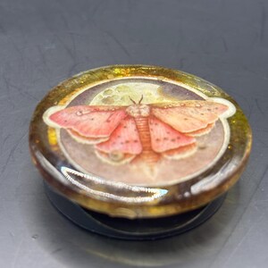 May include: A round, black and gold pop-up phone grip featuring a detailed illustration of a moth with pink and orange wings against a full moon. The grip has a clear, glossy finish.