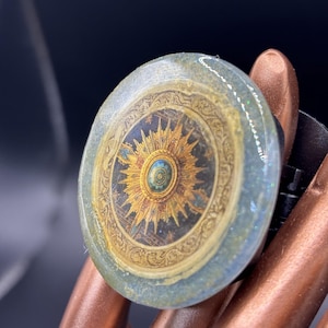 May include: A round ring with a detailed compass design. The ring features a gold and blue color scheme with intricate patterns. The ring is held by a person's fingers, and the background is dark.