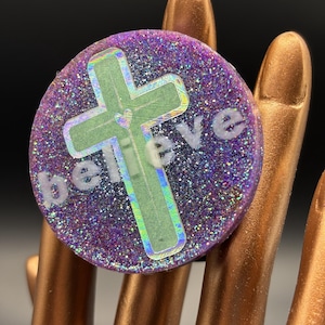 May include: A round, purple glittered accessory with the word "believe" in white lettering. A green cross is centered on the accessory. The accessory is held by a hand with gold-colored nails.