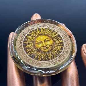 May include: A round ring with a smiling sun design. The ring features a detailed illustration of a sun with rays, set against a cream-colored background. The ring is set in a clear resin and has a dark brown base.
