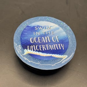 May include: A round, blue phone grip with the words "SWIM IN THE OCEAN OF UNCERTAINTY" in white. The design features a watercolor ocean scene with a glittery border and a black base.