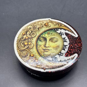May include: A round, decorative box featuring a celestial design. The lid showcases a detailed illustration of a serene face within a crescent moon, with gold and white accents. The rim is dark red with glitter.