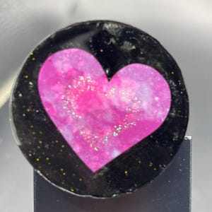 May include: A round, black resin heart-shaped phone grip with a pink and white marbled heart design. The black resin has gold glitter accents. The phone grip is mounted on a black stand.