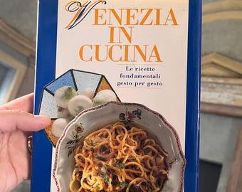 May include: A cookbook titled "Venezia in Cucina" with a blue spine and a photo of a plate of pasta on the cover. The text on the cover reads "Le ricette fondamentali gesto per gesto" and "Arnoldo Mondadori Editore".