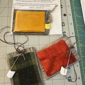 May include: Three handmade "Scratch Tools" for quilters. Each tool features a rectangular wooden frame with a fabric insert in shades of orange, green plaid, and red. A safety pin and twine are attached for hanging. The text on the packaging reads "Scratch Tool".