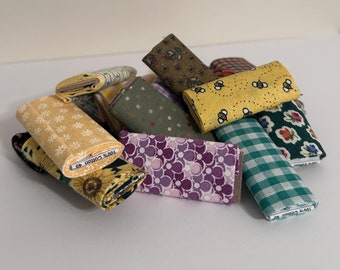 May include: A pile of fabric scraps in various patterns and colors, including yellow, green, purple, and white. The fabric scraps are folded and stacked on a white surface. The text "100% COTTON 42"