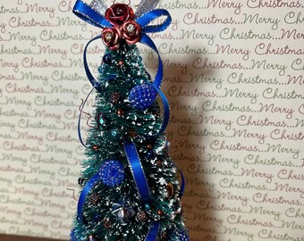 May include: A miniature Christmas tree decorated with blue and silver baubles, a blue ribbon with a red rose accent, and a white base with silver snowflake accents. The tree is surrounded by small, silver gift boxes.