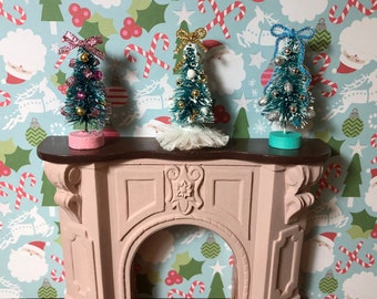 May include: A miniature light pink fireplace mantel with a dark brown top shelf. Three small bottle brush Christmas trees stand on the mantel, decorated with colorful beads and topped with bows in pink, gold, and blue, with a white ruffled base.