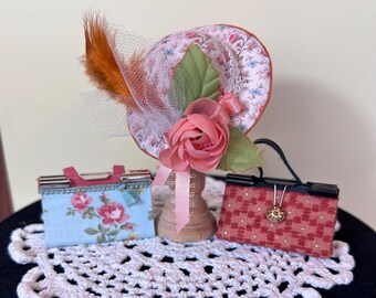 May include: Three miniature purses and a hat with a pink flower and a feather on a white crocheted doily. The items are on a black rotating display stand.
