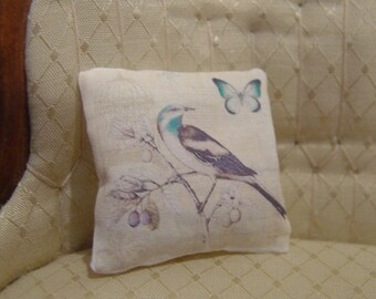May include: A white pillow with a floral design featuring a bird and a butterfly. The pillow is on a cream-colored couch with a diamond pattern.