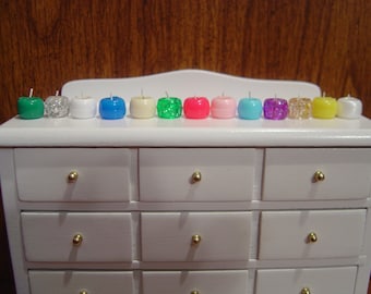 May include: A row of thirteen small, round, apple-shaped candles in various colors and finishes, including solid green, white, blue, yellow, pink, and light blue, alongside glittery, pearlescent, and translucent options, each featuring a single white wick.