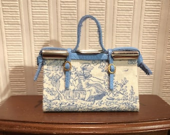 May include: A small blue and white patterned handbag with a silver clasp and gold accents. The bag has a top handle and a blue leather trim.