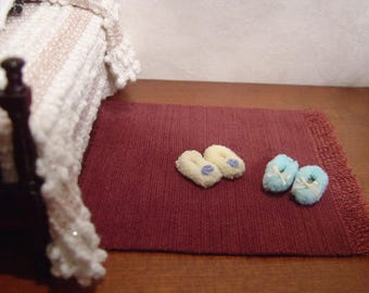 May include: Two pairs of fluffy slippers, one pair is light blue and the other is cream coloured. The slippers are on a burgundy rug with a white fringe.