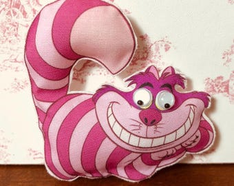May include: Pink and white striped fabric Cheshire Cat with a wide grin and large, white eyes. The cat is lying on its side with its tail curled around its body.