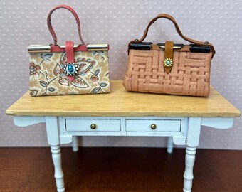 May include: Two miniature handbags on a wooden table. One handbag is pink with a woven pattern and the other is beige with a floral pattern. Both handbags have a brown leather handle and a metal clasp.