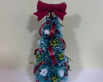 May include: A miniature Christmas tree with a teal base and branches. It is decorated with red ribbon, green ornaments, and white accents. A large red bow sits atop the tree, with small gold bells. The tree is approximately 6 inches tall.