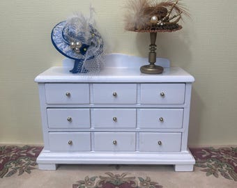 May include: A white dresser with 12 drawers and silver knobs. Two miniature hats are displayed on top of the dresser. One hat is brown with feathers and the other is blue with white feathers.
