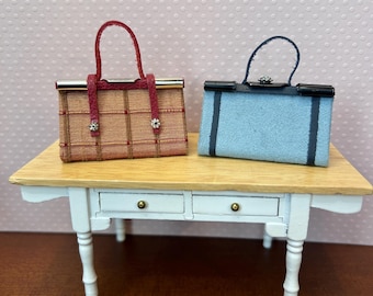 May include: Two miniature handbags on a white table with two drawers. The handbag on the left is brown with a red handle and gold accents. The handbag on the right is light blue with a black handle and gold accents.