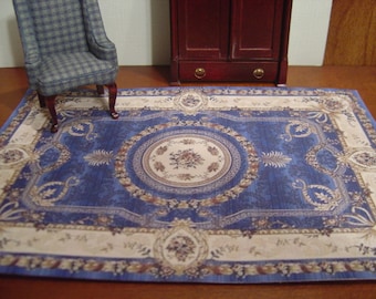 May include: A rectangular area rug featuring a deep blue center with a central cream-colored medallion containing floral motifs. The border displays intricate cream and tan scrollwork and floral patterns, creating a detailed, symmetrical design.