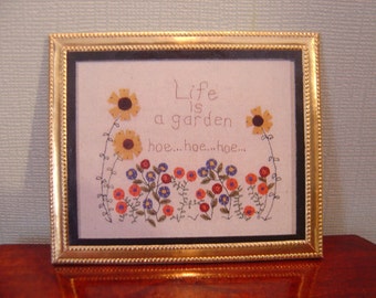 May include: A framed embroidery with a gold frame and a white background. The embroidery depicts a garden scene with flowers and the text "Life is a garden hoe...hoe...hoe..."