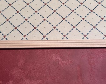 May include: A close-up of beige wallpaper with a repeating pattern of brown hearts and black dashed lines. The wallpaper is bordered by a thin, light brown trim. The bottom of the image shows a solid, dark red surface.